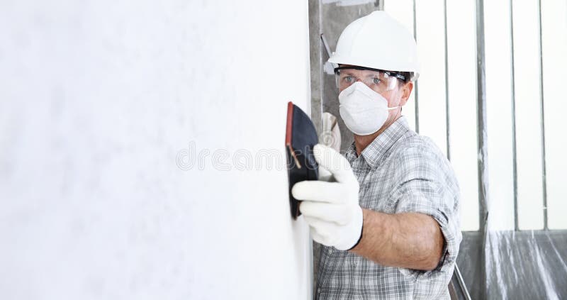 Man sand the wall with sandpaper, professional construction worker with mask, safety hard hat, gloves and protective glasses.