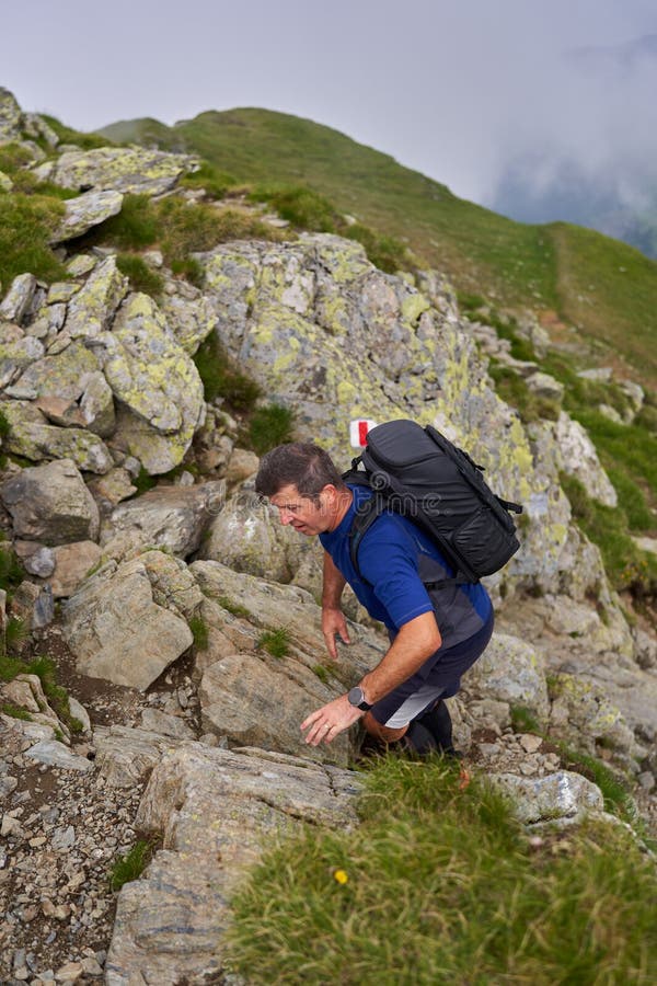 Man scrambling rocky mountain path with backpack up trail on overcast day imagens de stock