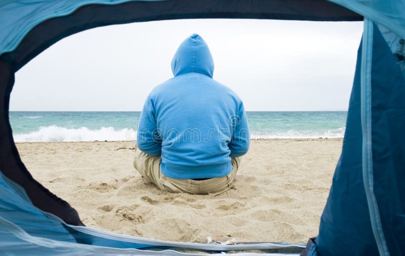 A man is sitting alone on the beach and looking out to sea as viewed through the inside of a tent. He is wearing a blue hooded sweatshirt and is camping on the beach. Serene beach atmosphere stock images, royalty-free photos and pictures