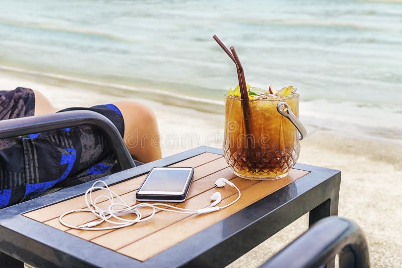 Man sitting at the beach with cocktail bucket, cell phone and earpods on table. Blurred sea in background. Vacation, travel