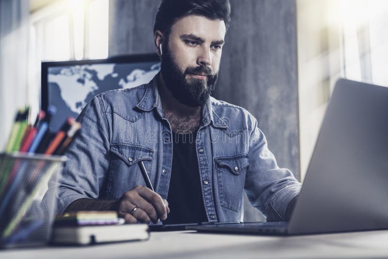 Young man sitting at window and working on laptop. Bearded man sitting at  desktop with smartphone in his hands. Online education. Stock Photo