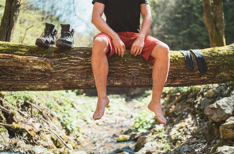 Man sitting on the fallen tree log over the mountain forest stream while he waiting for socks laundry drying and trekking boots.