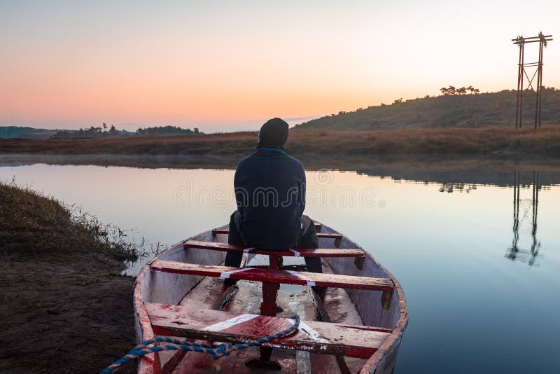 Man sitting at traditional wood boat at calm lake with dramatic colorful sky reflection at morning in details