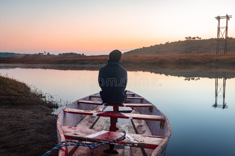 Man sitting at traditional wood boat at calm lake with dramatic colorful sky reflection at morning in details.