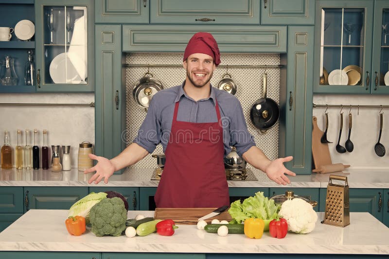 Man smile in chef hat in kitchen. Happy cook at table. Vegetables and tools ready for cooking dishes. Food preparation