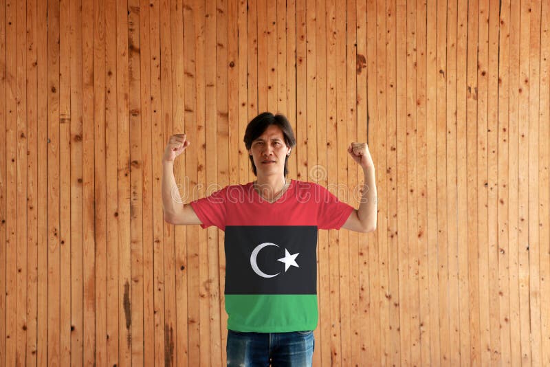 Man som bär Libyen flaggafärg av skjortan och står med den lyftta både näven på träväggbakgrunden royaltyfri bild Man som bär Libyen flaggafärg av skjortan och står med den lyftta både näven på träväggbakgrunden royaltyfri bild