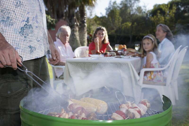 Man som grillar med familjen på den utomhus- tabellen royaltyfri foto Man som grillar med familjen på den utomhus- tabellen royaltyfri foto