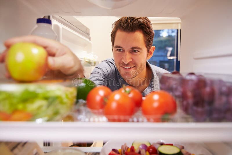 Man som mycket ser den inre kylen av mat och väljer Apple royaltyfri bild Man som mycket ser den inre kylen av mat och väljer Apple royaltyfri bild