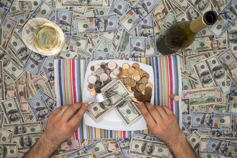 Conceptual image of a man eating money through extravagance with an overhead view of him sitting at a table covered in 100 dollar bills eating money on a plate with a glass and bottle of champagne. Conceptual image of a man eating money through extravagance with an overhead view of him sitting at a table covered in 100 dollar bills eating money on a plate with a glass and bottle of champagne