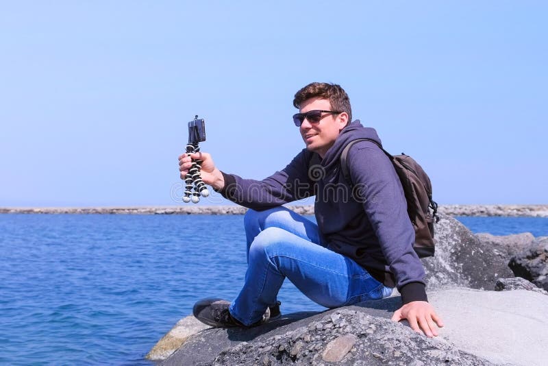 Man spricht über Video-Chat-Telefon mit kleinem Stativ kommt zum Meer Strand sitzt auf Stein lizenzfreie stockfotografie