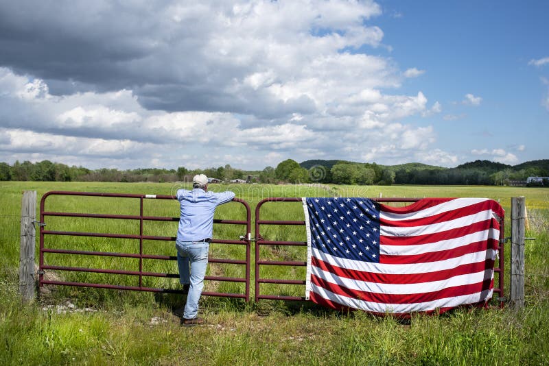 Man standing at metal gate in beautiful grass field, American flag hanging on fence