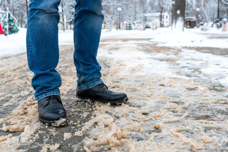 A man stands on the sidewalk in snow and mud. Black shoes and blue jeans close-up on the background of dirty snow. Ice on the road