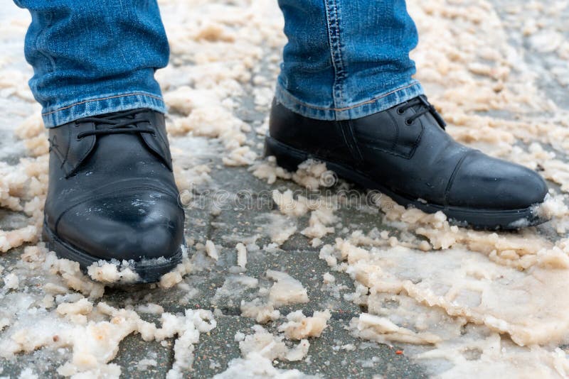 A man stands on the sidewalk in snow and mud. Black shoes and blue jeans close-up on the background of dirty snow. Ice on the road