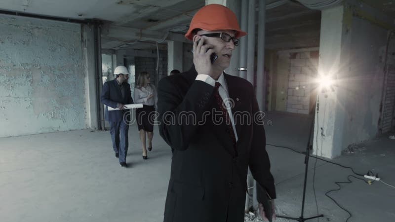 Man in suit, helmet call inside construction site. At background team of young builders man, african