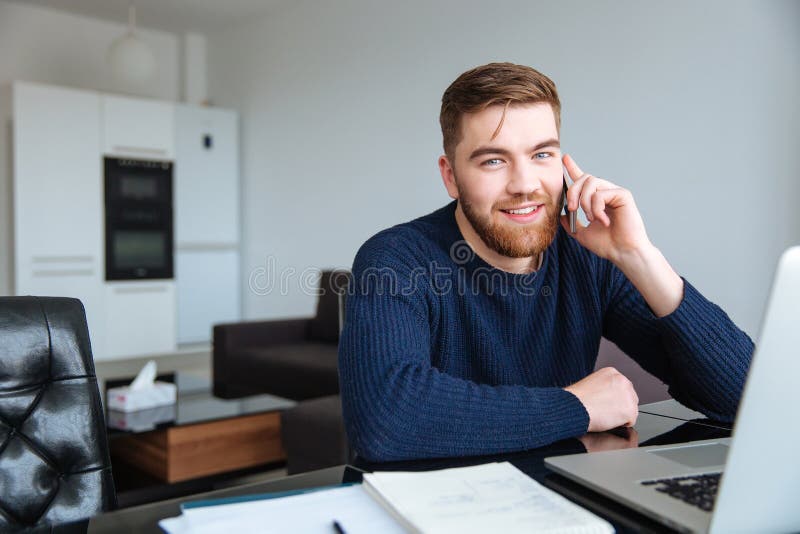 Portrait of a smiling man sitting at the table with laptop and talking on the phone at home. Looking at camera. Portrait of a smiling man sitting at the table with laptop and talking on the phone at home. Looking at camera