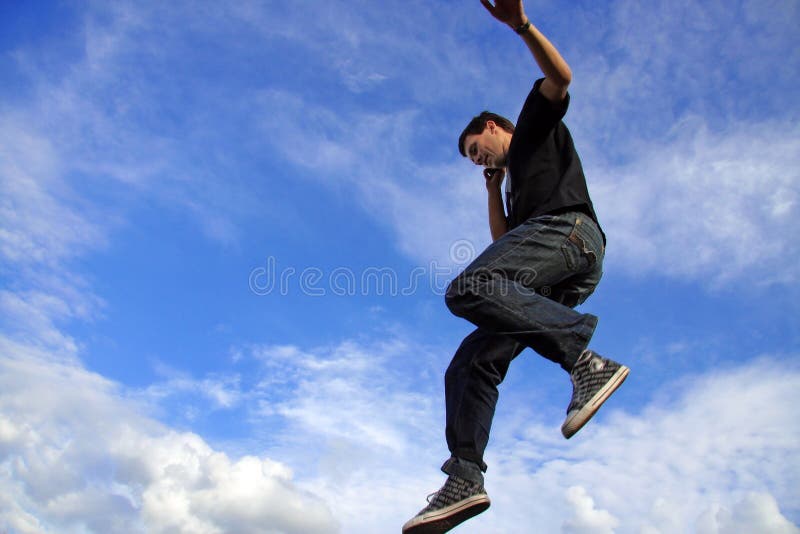 Low angle side view of young man jumping in air and talking on mobile telephone, blue sky and cloudscape background. Low angle side view of young man jumping in air and talking on mobile telephone, blue sky and cloudscape background.