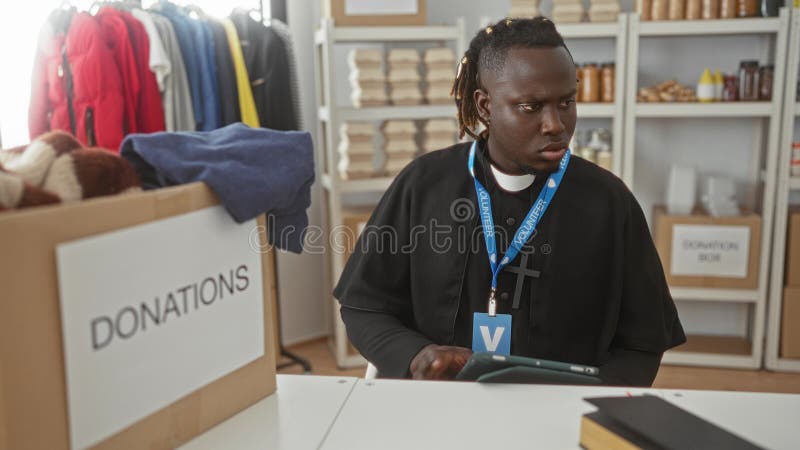 Man tapping tablet and sorting clothes donations at a donations desk inside a volunteer center building with labeled boxes and shelves; compassion service. Charity sorting stock images, royalty-free photos and pictures