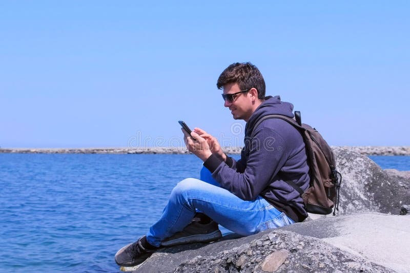 Man taveller Chat-Typ-Nachricht auf Smartphone sitzt auf Stein am Strand stockfoto