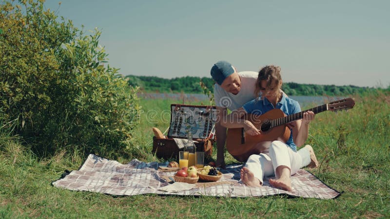 A man teaches a woman to play the guitar at a picnic in the summer.