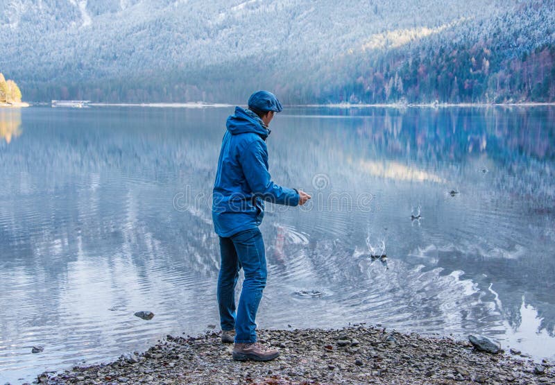 A man is throwing a little stone in the lake registering the motion of the splashes in the water. The landscape is peaceful and quiet. Man dressed with blue jeans and blue jacket in a blue landscape. Motion photography. A man is throwing a little stone in the lake registering the motion of the splashes in the water. The landscape is peaceful and quiet. Man dressed with blue jeans and blue jacket in a blue landscape. Motion photography.