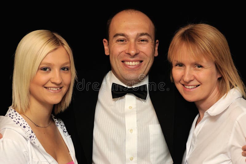 A studio portrait of a smiling man wearing a suit and bow tie with his arms around two pretty women on either side. Black background. A studio portrait of a smiling man wearing a suit and bow tie with his arms around two pretty women on either side. Black background.