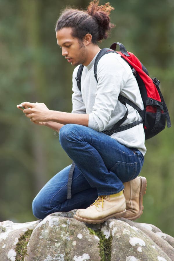 Man Using Mobile Phone Whilst Hiking In Countryside Kneeling Down Wearing A Backpack