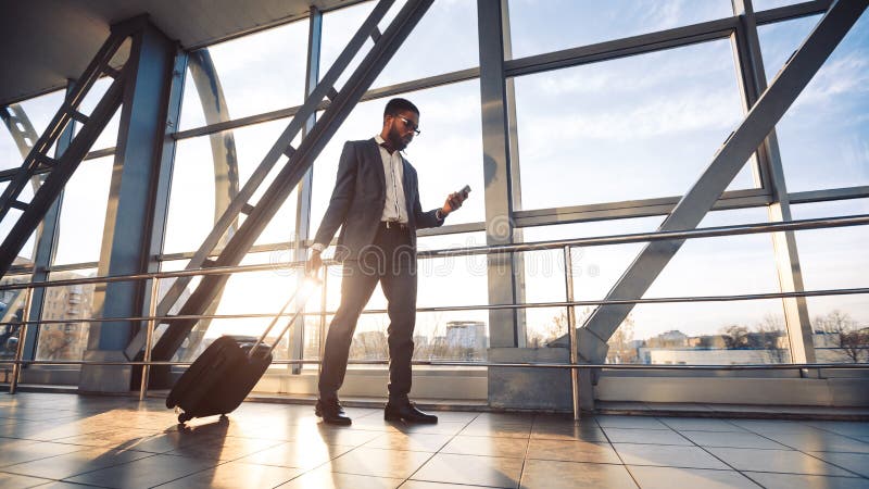 Black Man Using Phone And Business Trip App Walking With Suitcase To Gate In Airport Terminal. Panorama, Free Space. Black Man Using Phone And Business Trip App Walking With Suitcase To Gate In Airport Terminal. Panorama, Free Space