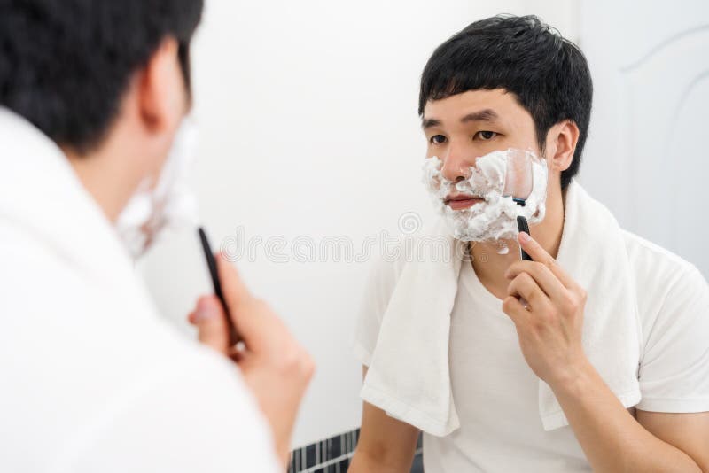 Man Using Razor To Shaving His Face with Cream Foam in Bathroom Mirror