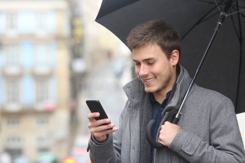 Portrait of a handsome man using a smart phone in a rainy day holding an umbrella in the street