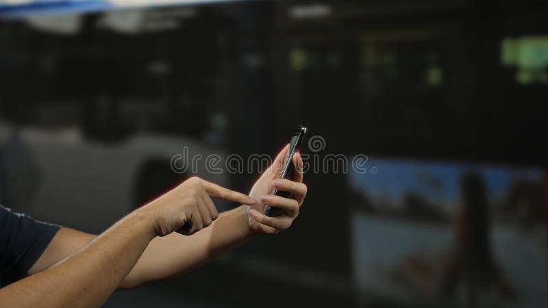 Man using smartphone outdoors at bus stop, focused on hands interacting with screen against blurred background, emphasizing stock images