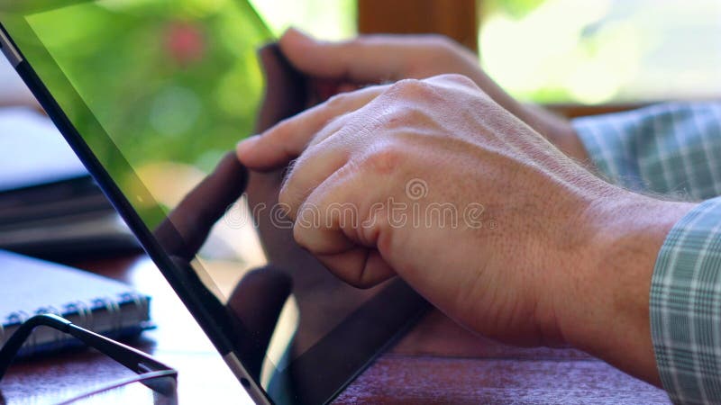 Man using tablet device closeup at desk in front of window. Man using tablet device closeup at desk, backlit by sunshine with reflections through window.