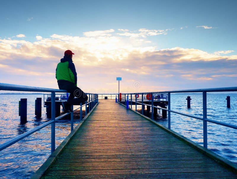 Man in warm jacket and baseball cap sit on pier and enjoy quiet morning sea. Tourist relax. royalty free stock photography Man in warm jacket and baseball cap sit on pier and enjoy quiet morning sea. Tourist relax. royalty free stock photography