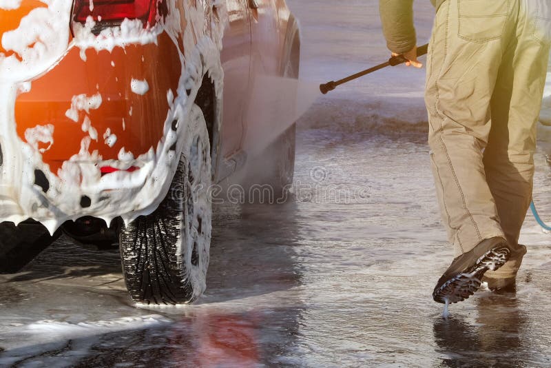 Man washes his orange car at car wash on clear sunny day. Cleaning with soap suds at self-service car wash