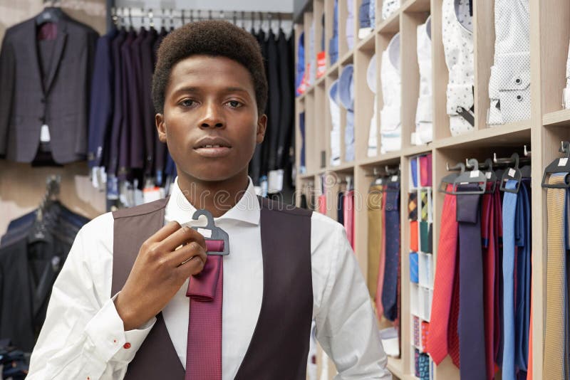 Young client choosing and holding tie. Man wearing in white shirt and black waistcoat posing and looking at camera. Many elegant clothing for man in fashionable boutique. Young client choosing and holding tie. Man wearing in white shirt and black waistcoat posing and looking at camera. Many elegant clothing for man in fashionable boutique.
