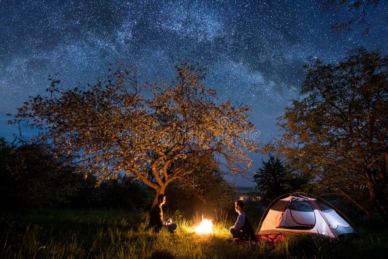 Man and women tourists sitting at a campfire near tent under trees and beautiful night sky full of stars and milky way. Night camping. Astrophotography. Man and women tourists sitting at a campfire near tent under trees and beautiful night sky full of stars and milky way. Night camping. Astrophotography