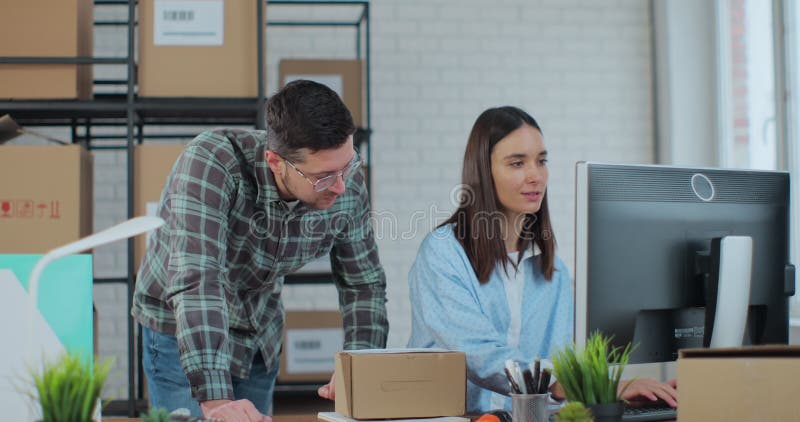 A man and a woman work in a warehouse. Young woman using computer, man confirming contact details on boxes. stock video footage