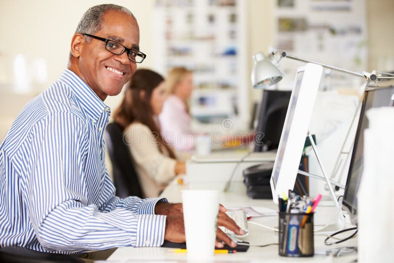 Man Working At Desk In Busy Creative Office Smiling