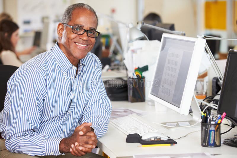 Man Working At Desk In Busy Creative Office Smiling