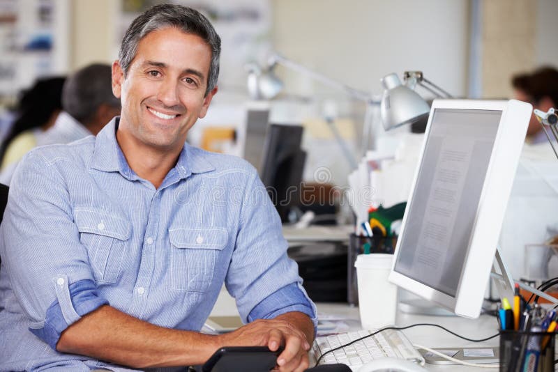 Man Working At Desk In Busy Creative Office Smiling