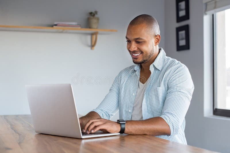 Young casual man working on laptop at home. African businessman typing on computer to send an email. Happy smiling black man surfing the net in a living room.