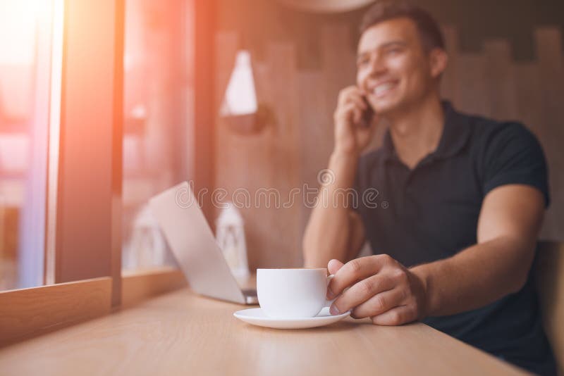 Focus on white cup. man working on net-book in a cafe, flare sun. Male talking on smart phone on background and holding a cup of coffee in foreground. Successful and rich person