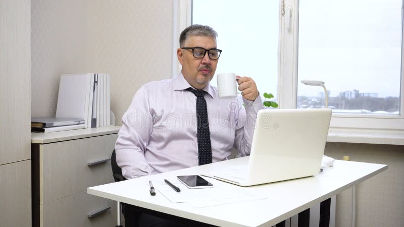 A man works at a computer, then looks contentedly at the screen and drinks coffee