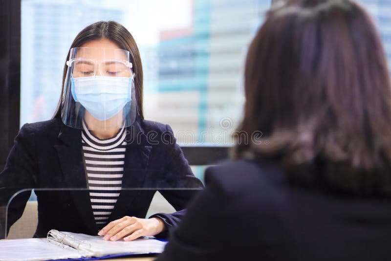 Manager from HR department wearing facial mask is interviewing new applicant and reading her resume and profile through the partition for social distancing and new normal policy. Manager from HR department wearing facial mask is interviewing new applicant and reading her resume and profile through the partition for social distancing and new normal policy