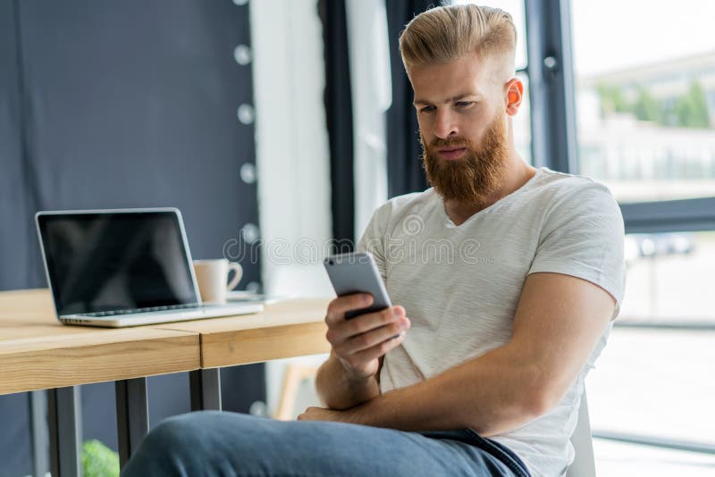 Mandando un sms al collega Giovane sicuro in Smart Phone astuto della tenuta di abbigliamento casual ed esaminarlo mentre sedendo immagini stock Mandando un sms al collega Giovane sicuro in Smart Phone astuto della tenuta di abbigliamento casual ed esaminarlo mentre sedendo immagini stock
