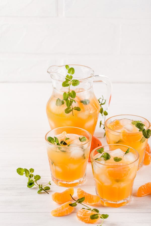 Mandarin cocktail with ice and mint in beautiful glasses and jug, fresh ripe citrus on white wooden background. Sweet orange juice