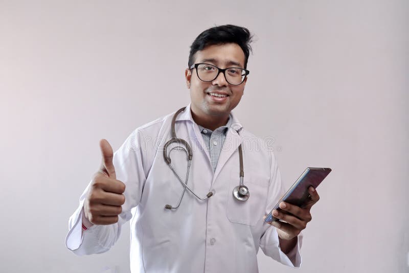 Manlig indianläkare i vit rock och stetoskop som visar tummen upp med smart telefon royaltyfria bilder Manlig indianläkare i vit rock och stetoskop som visar tummen upp med smart telefon royaltyfria bilder