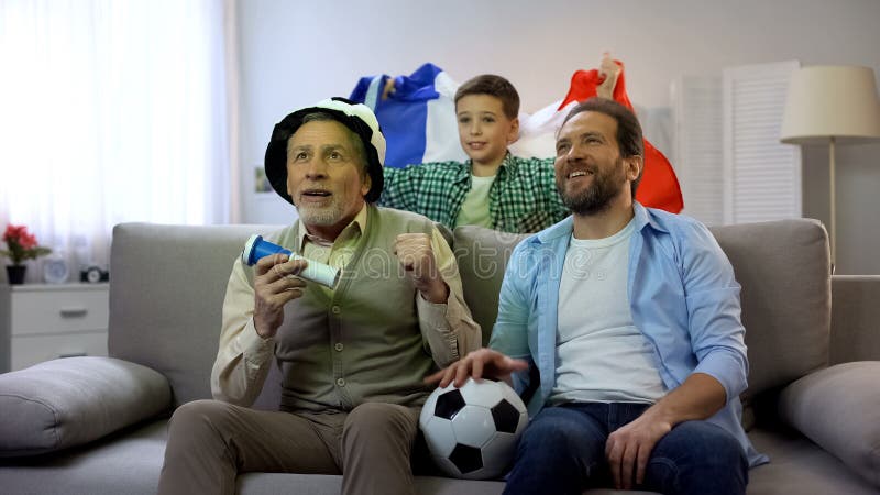 Manliga familjemedlemmar som hemma stöttar fransk fotboll, hållande ögonen på mästerskap royaltyfri fotografi Manliga familjemedlemmar som hemma stöttar fransk fotboll, hållande ögonen på mästerskap royaltyfri fotografi