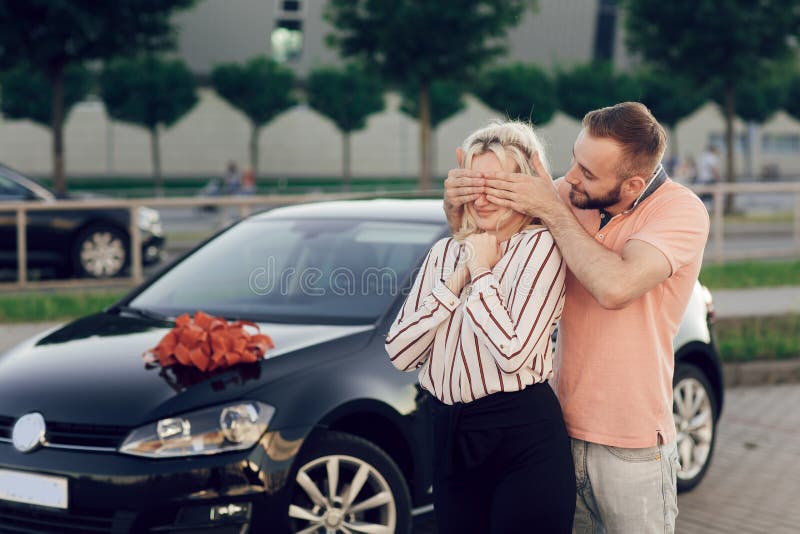 Mann, der frau durch kauf eines neuen autos überrascht. junges paar kauft  ein auto, mann und frau stehen in der nähe des autos auf der straße.  überraschungsgeschenk für einen geliebten menschen.