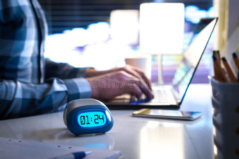 Man working late. Workaholic or being behind schedule concept. Business person in modern office building or home at night using laptop. Time in digital clock on table in workstation. Man working late. Workaholic or being behind schedule concept. Business person in modern office building or home at night using laptop. Time in digital clock on table in workstation.