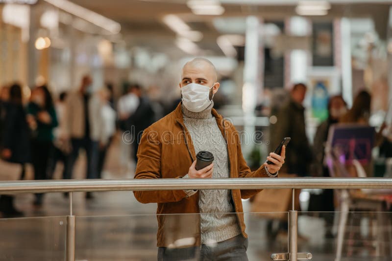 Mann in einer Gesichtsmaske benutzt ein Telefon und hält einen Kaffee im Einkaufszentrum lizenzfreie stockfotos